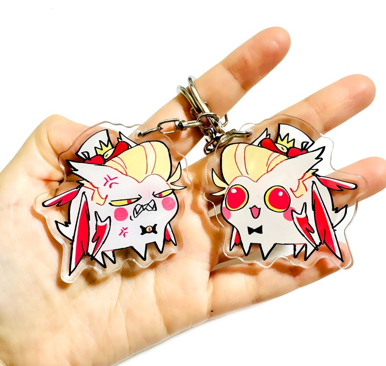 May include: Two clear acrylic keychains featuring a cartoon character with yellow hair, red wings, and a bow tie. One keychain shows the character with a surprised expression and the other shows the character with an angry expression.