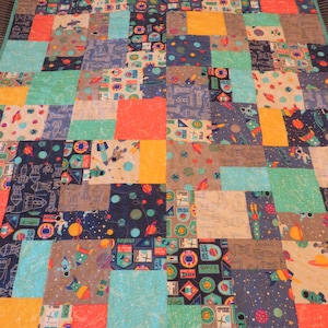 May include: A patchwork quilt with a space theme. The quilt is made of various fabrics with different space-themed patterns, including rockets, planets, stars, and astronauts. The quilt is in shades of blue, green, orange, and yellow.