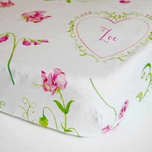 May include: White cot sheet with a pink floral pattern and a heart with the name "Zoe" in pink lettering.