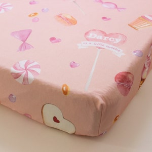 May include: Pink fitted cot sheet with a watercolour design of sweets, hearts, and cupcakes. The text "Darcy is a little sweetie" is printed on a heart-shaped lollipop.