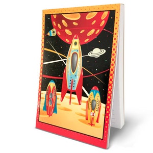 May include: A notebook with a colorful space-themed illustration. The cover features rockets, planets, and a starry background. The design includes red, yellow, blue, and orange hues, with a red border.