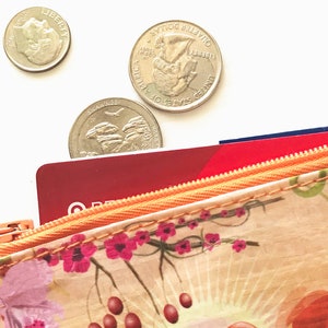 May include: Close-up of a floral patterned wallet with a zipper, partially open to reveal red and blue cards. Three US coins, a dime and two quarters, are scattered above the wallet on a white background. The wallet features pink flowers and red berries.