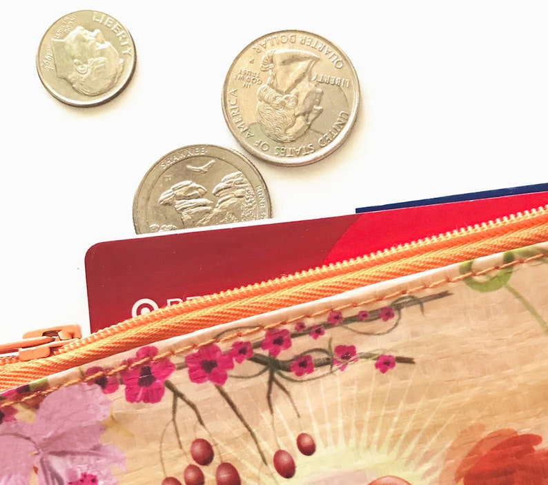 May include: Close-up of a floral patterned wallet with a zipper, partially open to reveal red and blue cards. Three US coins, a dime and two quarters, are scattered above the wallet on a white background. The wallet features pink flowers and red berries.
