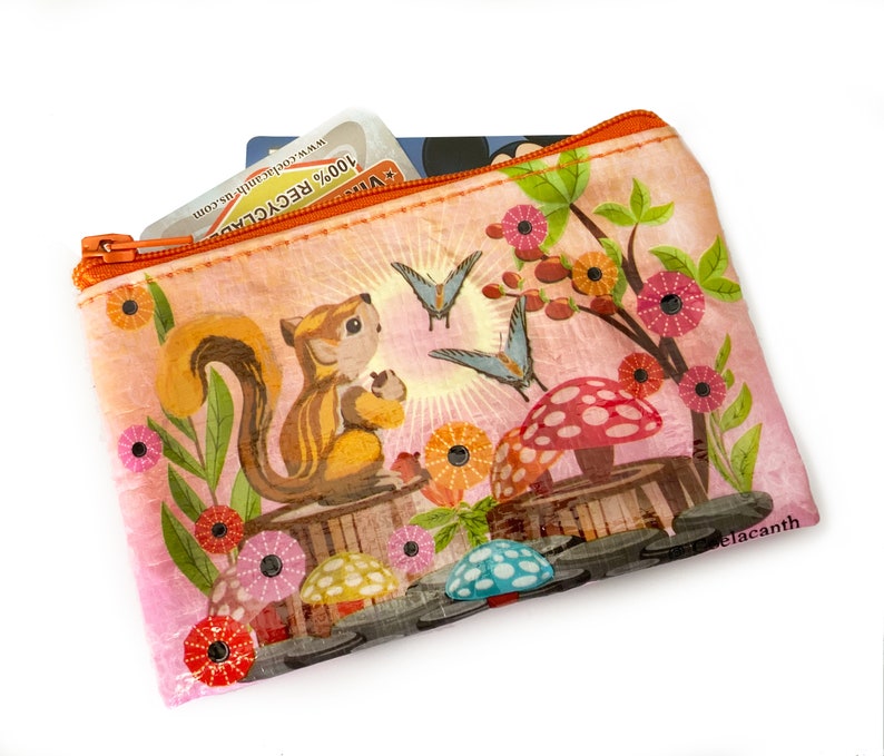 May include: A rectangular, zippered pouch with a whimsical illustration. The design features a squirrel, butterflies, mushrooms, and flowers in shades of orange, pink, and green. The pouch contains cards and has the text "100% RECYCLED".