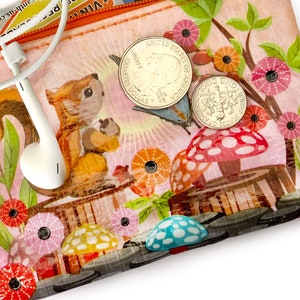 May include: A clear, zippered pouch with a whimsical design featuring a squirrel, mushrooms, and flowers. The pouch contains a pair of white earbuds and two US coins: a quarter and a dime. The background is a light pink with a forest theme.