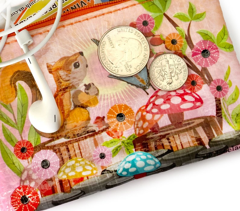 May include: A clear, zippered pouch with a whimsical design featuring a squirrel, mushrooms, and flowers. The pouch contains a pair of white earbuds and two US coins: a quarter and a dime. The background is a light pink with a forest theme.