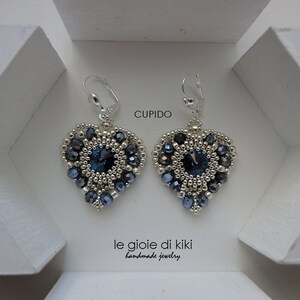Może przedstawiać: Para srebrnych kolczyk&oacute;w w kształcie serca z niebieskimi kryształowymi akcentami. Kolczyki są oznaczone "Cupido" i "le gioie di kiki handmade jewelry".