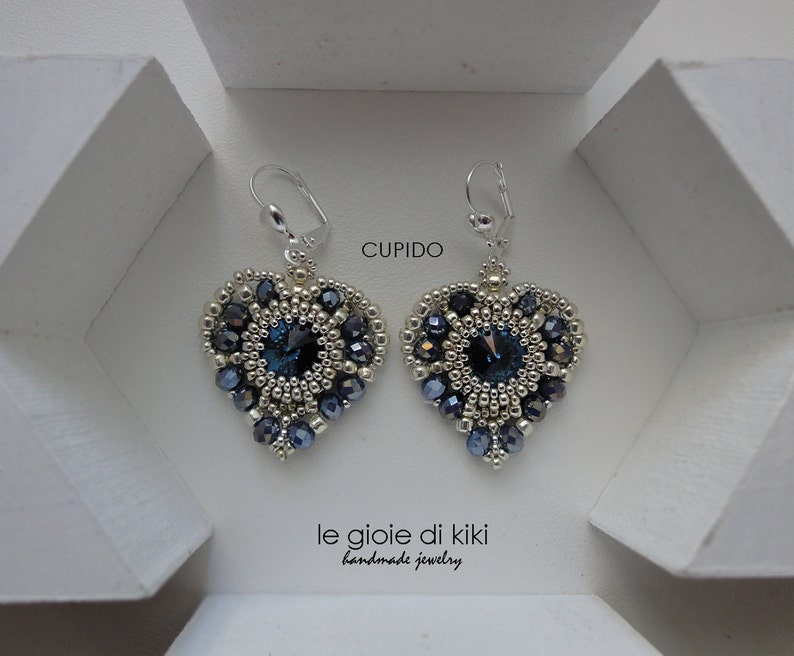 Może przedstawiać: Para srebrnych kolczyk&oacute;w w kształcie serca z niebieskimi kryształowymi akcentami. Kolczyki są oznaczone "Cupido" i "le gioie di kiki handmade jewelry".
