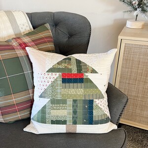 May include: A white pillow with a green and red patchwork Christmas tree design. The pillow is on a gray armchair with a plaid pillow beside it.
