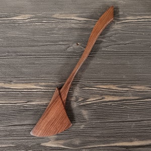 May include: A wooden spatula with a long, thin handle and a wide, flat blade. The spatula is made of dark wood and has a natural finish.
