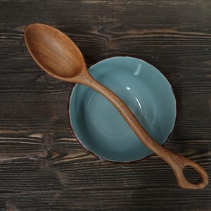 Op de afbeelding: Een houten lepel met een afgeronde kom en een handvat met een gat rust op een lichtblauwe kom. De lepel en kom liggen op een donker houten oppervlak. De lepel is gemaakt van lichtbruin hout.
