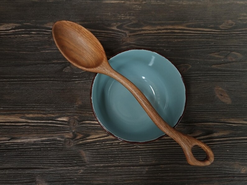 Op de afbeelding: Een houten lepel met een afgeronde kom en een handvat met een gat rust op een lichtblauwe kom. De lepel en kom liggen op een donker houten oppervlak. De lepel is gemaakt van lichtbruin hout.