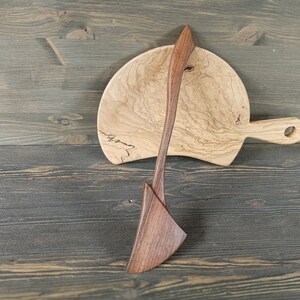 May include: A wooden serving spoon and a wooden cutting board with a handle. The cutting board is light brown with dark brown wood grain. The serving spoon is dark brown with a light brown handle.