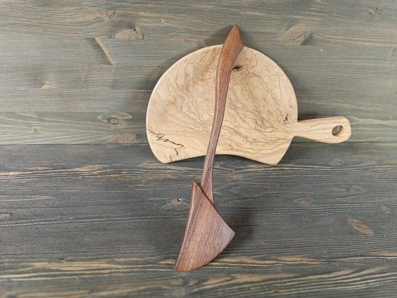 May include: A wooden serving spoon and a wooden cutting board with a handle. The cutting board is light brown with dark brown wood grain. The serving spoon is dark brown with a light brown handle.