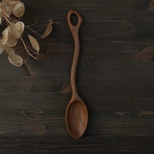 Op de afbeelding: Een handgemaakte houten lepel met een lange, gebogen steel en een afgeronde kom. De lepel is gemaakt van donker hout en heeft een gat aan het uiteinde van de steel. De lepel wordt getoond op een donker houten oppervlak.