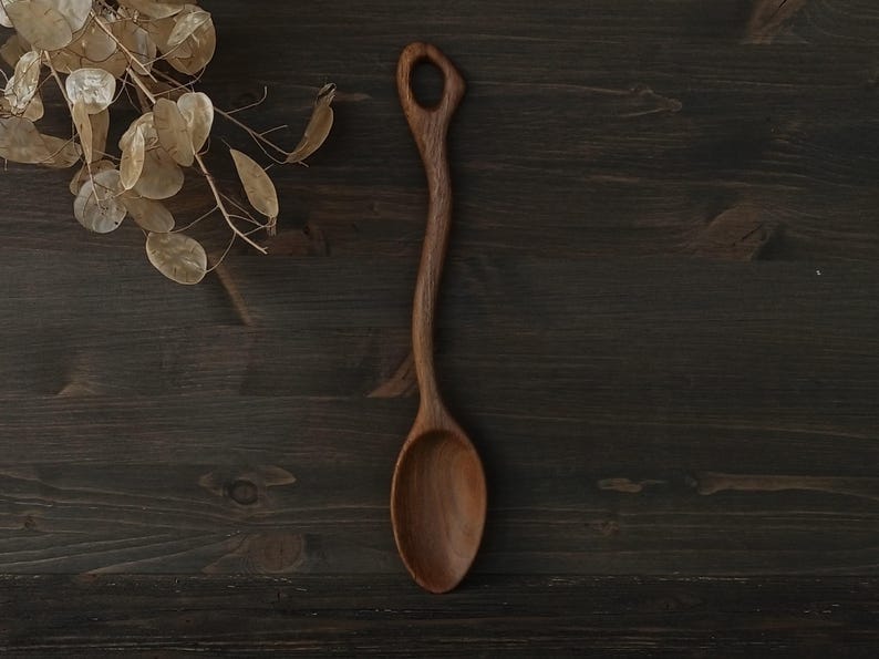 Op de afbeelding: Een handgemaakte houten lepel met een lange, gebogen steel en een afgeronde kom. De lepel is gemaakt van donker hout en heeft een gat aan het uiteinde van de steel. De lepel wordt getoond op een donker houten oppervlak.