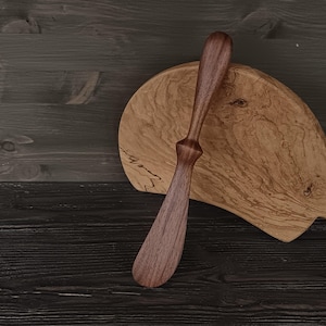 May include: A dark brown wooden butter spreader with a smooth, rounded handle and a wide, flat blade, positioned in front of a light-coloured, semi-circular wooden cutting board featuring a prominent natural wood grain pattern and texture.