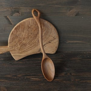 Op de afbeelding: Een houten serveerlepel en een snijplank. De lepel heeft een lange steel en een afgeronde kom. De snijplank is ovaal van vorm met een handvat. Beide items zijn gemaakt van lichtgekleurd hout.