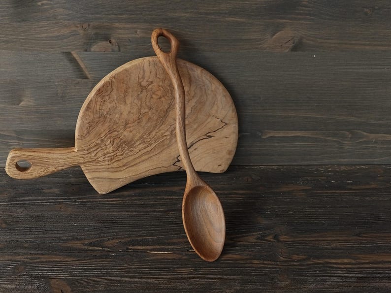 Op de afbeelding: Een houten serveerlepel en een snijplank. De lepel heeft een lange steel en een afgeronde kom. De snijplank is ovaal van vorm met een handvat. Beide items zijn gemaakt van lichtgekleurd hout.
