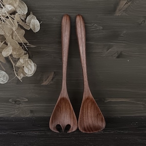 Könnte beinhalten: Zwei Salatbestecke aus Holz mit natürlicher Holzmaserung. Eines der Besteckteile hat ein geschlitztes Design, das andere ist massiv. Sie werden auf einer dunklen Holzoberfläche präsentiert, was auf eine rustikale oder ländliche Ästhetik hindeutet.