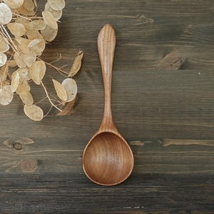 Könnte beinhalten: Ein Holzlöffel mit einem abgerundeten Löffel und einem langen, schlanken Griff. Der Löffel ist aus hellbraunem Holz gefertigt und liegt auf einer dunkelbraunen Holzoberfläche. Getrocknete, beigefarbene Blätter befinden sich in der oberen linken Ecke.