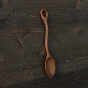 Op de afbeelding: Een handgemaakte houten lepel met een lange, slanke steel en een afgeronde kom. De lepel is gemaakt van lichtbruin hout en heeft een gat aan het uiteinde van de steel. De lepel wordt gepresenteerd tegen een donkere houten achtergrond.