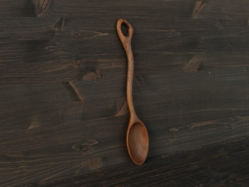 Op de afbeelding: Een handgemaakte houten lepel met een lange, slanke steel en een afgeronde kom. De lepel is gemaakt van lichtbruin hout en heeft een gat aan het uiteinde van de steel. De lepel wordt gepresenteerd tegen een donkere houten achtergrond.