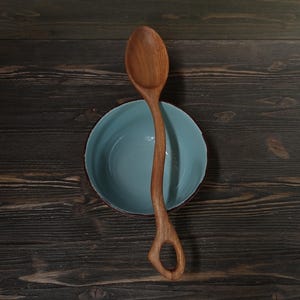 Op de afbeelding: Een houten lepel met een uniek handvatontwerp ligt over een lichtblauwe kom met een rode rand. De lepel is gemaakt van lichtbruin hout en heeft een gat aan het einde van het handvat. De kom staat op een donker houten oppervlak.