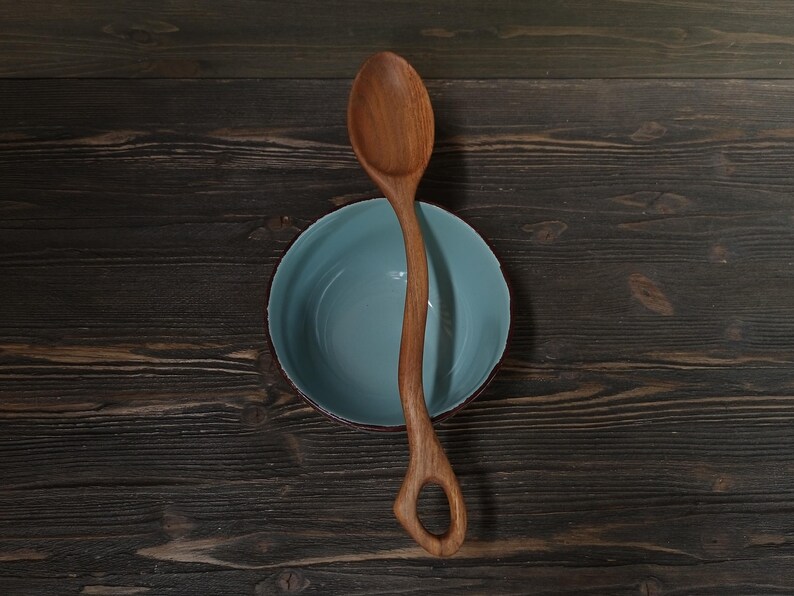 Op de afbeelding: Een houten lepel met een uniek handvatontwerp ligt over een lichtblauwe kom met een rode rand. De lepel is gemaakt van lichtbruin hout en heeft een gat aan het einde van het handvat. De kom staat op een donker houten oppervlak.