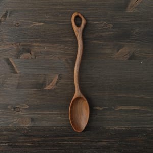 Op de afbeelding: Een handgemaakte houten lepel met een lange, gebogen steel en een afgeronde kom. De lepel is gemaakt van lichtbruin hout en heeft een gat aan het uiteinde van de steel. De lepel is ongeveer 30 cm lang.
