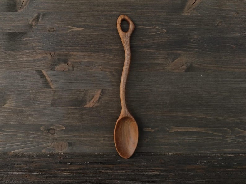 Op de afbeelding: Een handgemaakte houten lepel met een lange, gebogen steel en een afgeronde kom. De lepel is gemaakt van lichtbruin hout en heeft een gat aan het uiteinde van de steel. De lepel is ongeveer 30 cm lang.