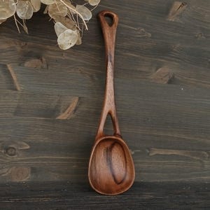 May include: A handcrafted wooden spoon with a long handle and a rounded bowl. The spoon is made of dark brown wood and features a unique cutout design near the bowl. The spoon is approximately 12 inches long.