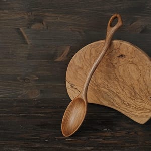 Op de afbeelding: Een handgemaakte houten lepel met een gebogen handvat en een afgeronde kom rust op een houten snijplank. De lepel is lichtbruin en de plank heeft een natuurlijk houtnerfpatroon. De achtergrond is een donkerbruin houten oppervlak.