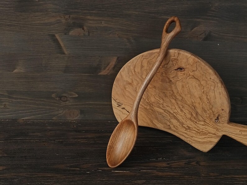 Op de afbeelding: Een handgemaakte houten lepel met een gebogen handvat en een afgeronde kom rust op een houten snijplank. De lepel is lichtbruin en de plank heeft een natuurlijk houtnerfpatroon. De achtergrond is een donkerbruin houten oppervlak.