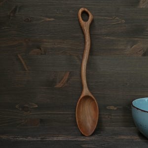 Op de afbeelding: Een handgemaakte houten lepel met een lange, gebogen steel en een afgeronde kom. De lepel is gemaakt van lichtbruin hout en heeft een gat aan het einde van de steel. De lepel ligt op een donkerbruin houten oppervlak.