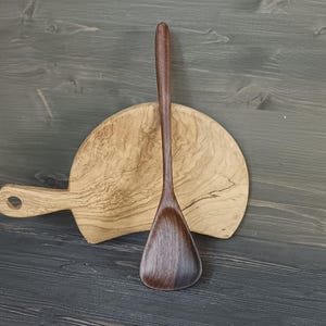 Könnte beinhalten: Ein hölzerner Kochlöffel mit breitem, flachem Kopf und langem Stiel liegt auf einem runden Holzschneidebrett mit Griff. Der Löffel und das Brett sind aus hellem und dunklem braunem Holz gefertigt.