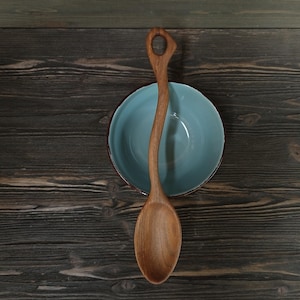 Op de afbeelding: Een houten lepel met een gebogen handvat en een afgeronde kom rust over een lichtblauwe kom. De lepel is gemaakt van lichtbruin hout en heeft een gat aan het einde van het handvat. De kom heeft een donkere rand en staat tegen een donkere houten achtergrond.