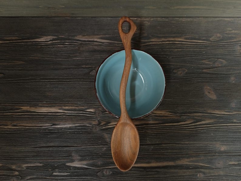 Op de afbeelding: Een houten lepel met een gebogen handvat en een afgeronde kom rust over een lichtblauwe kom. De lepel is gemaakt van lichtbruin hout en heeft een gat aan het einde van het handvat. De kom heeft een donkere rand en staat tegen een donkere houten achtergrond.
