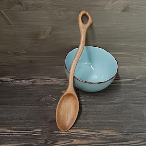 Op de afbeelding: Een houten lepel met een gebogen handvat en een ronde kom ligt naast een lichtblauwe kom met een bruine rand. De lepel is gemaakt van lichtgekleurd hout en heeft een gat aan het einde van het handvat. De kom is leeg.