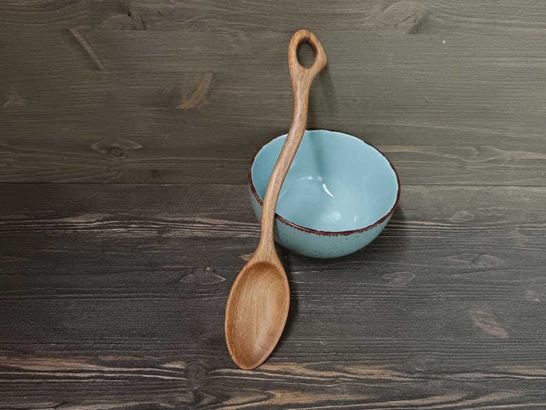 Op de afbeelding: Een houten lepel met een gebogen handvat en een ronde kom ligt naast een lichtblauwe kom met een bruine rand. De lepel is gemaakt van lichtgekleurd hout en heeft een gat aan het einde van het handvat. De kom is leeg.