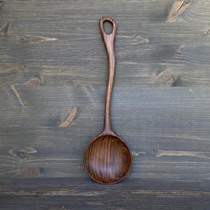 May include: A handcrafted wooden spoon with a long, curved handle and a round bowl. The spoon is made of dark brown wood and has a hole at the end of the handle for hanging. The spoon is displayed on a wooden surface.