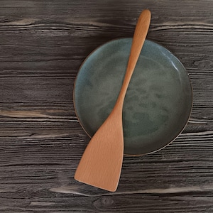 May include: A wooden spatula with a light brown finish sits on a dark green ceramic plate. The plate and spatula are on a dark wood surface.