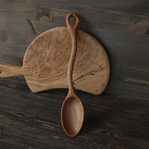 Op de afbeelding: Een houten lepel met een lange, gebogen steel en een afgeronde kom ligt op een houten snijplank. De lepel heeft een gat aan het uiteinde van de steel. De plank heeft een handvat en een natuurlijk houtnerfpatroon. De achtergrond is een donker houten oppervlak.
