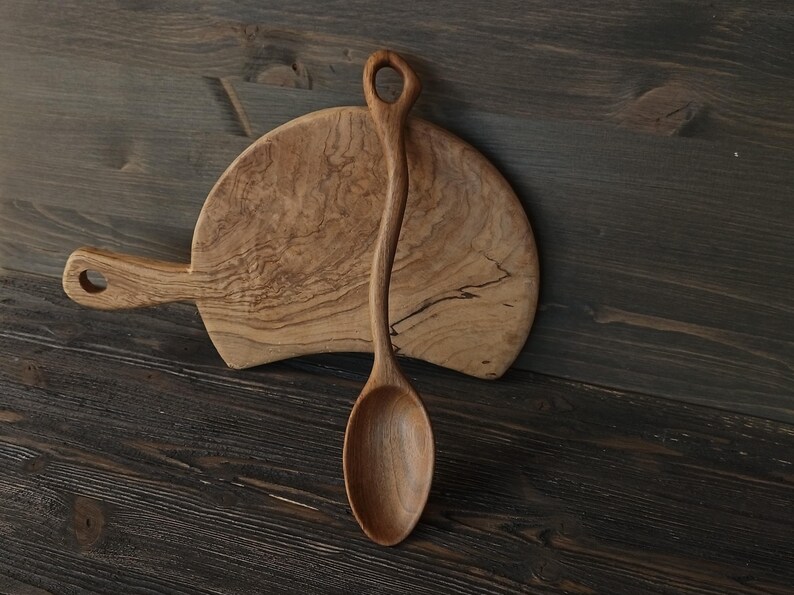 Op de afbeelding: Een houten lepel met een lange, gebogen steel en een afgeronde kom ligt op een houten snijplank. De lepel heeft een gat aan het uiteinde van de steel. De plank heeft een handvat en een natuurlijk houtnerfpatroon. De achtergrond is een donker houten oppervlak.