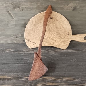 May include: A wooden serving spoon and a wooden cutting board with a natural wood grain pattern. The spoon is made of dark wood and the board is made of light wood.