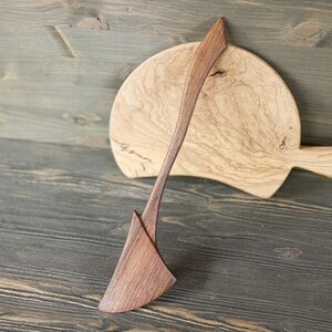May include: A wooden spatula with a curved handle and a wide, flat blade. The spatula is made of dark wood and has a natural finish. It is resting on a wooden cutting board with a light brown finish.