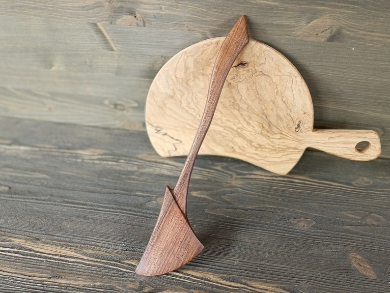 May include: A wooden spatula with a curved handle and a wide, flat blade. The spatula is made of dark wood and has a natural finish. It is resting on a wooden cutting board with a light brown finish.