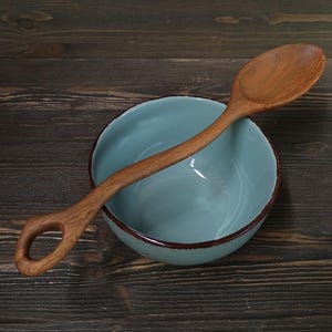 Op de afbeelding: Een houten lepel met een gebogen handvat en een gat aan het uiteinde rust op een lichtblauwe kom met een bruine rand. De lepel en kom staan op een donkere houten ondergrond.