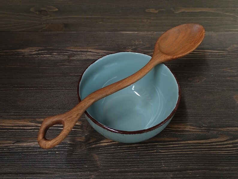 Op de afbeelding: Een houten lepel met een gebogen handvat en een gat aan het uiteinde rust op een lichtblauwe kom met een bruine rand. De lepel en kom staan op een donkere houten ondergrond.