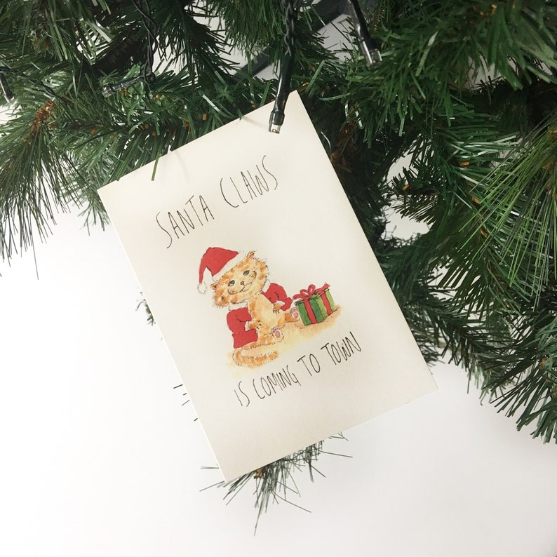Puede incluir: Una tarjeta blanca con el texto "Santa Claus is coming to town!" escrito en marcador negro. Una ilustraci&oacute;n de dibujos animados de un gato vestido de Pap&aacute; Noel sosteniendo un regalo envuelto est&aacute; en la tarjeta.